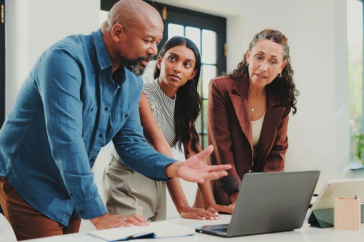 A group of senior IT employees gathers around a laptop discussing Microsoft’s new Teams licensing updates, recognizing how the simplified structure, broader feature access, and clearer SKUs will help them deploy collaboration tools more efficiently across their organization.