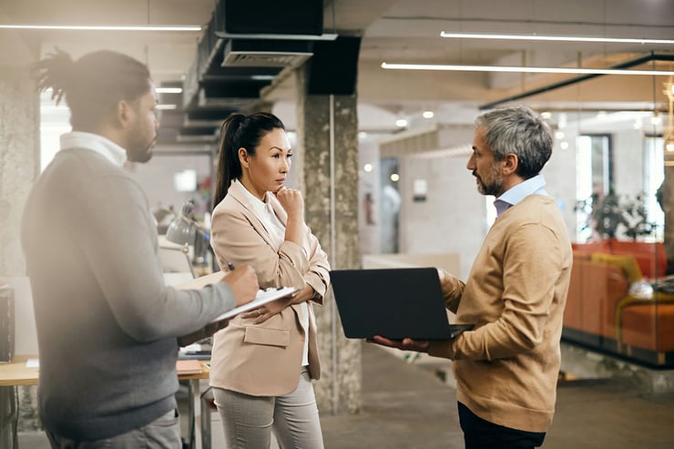 Three employees stand in a breakout area discussing how adopting a practical AI strategy for business—like using Copilot for drafting and data insights—can streamline their workflows and boost organizational productivity.