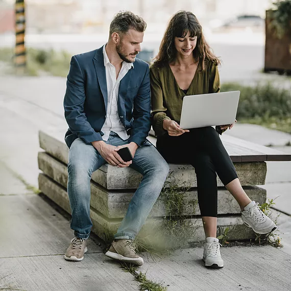 On their company campus, a male and female employee from a financial services organization collaborate outside, reviewing a PowerPoint deck on a work computer powered by Microsoft 365, refining their slides in real time to ensure a polished and professional presentation before meeting with a client.